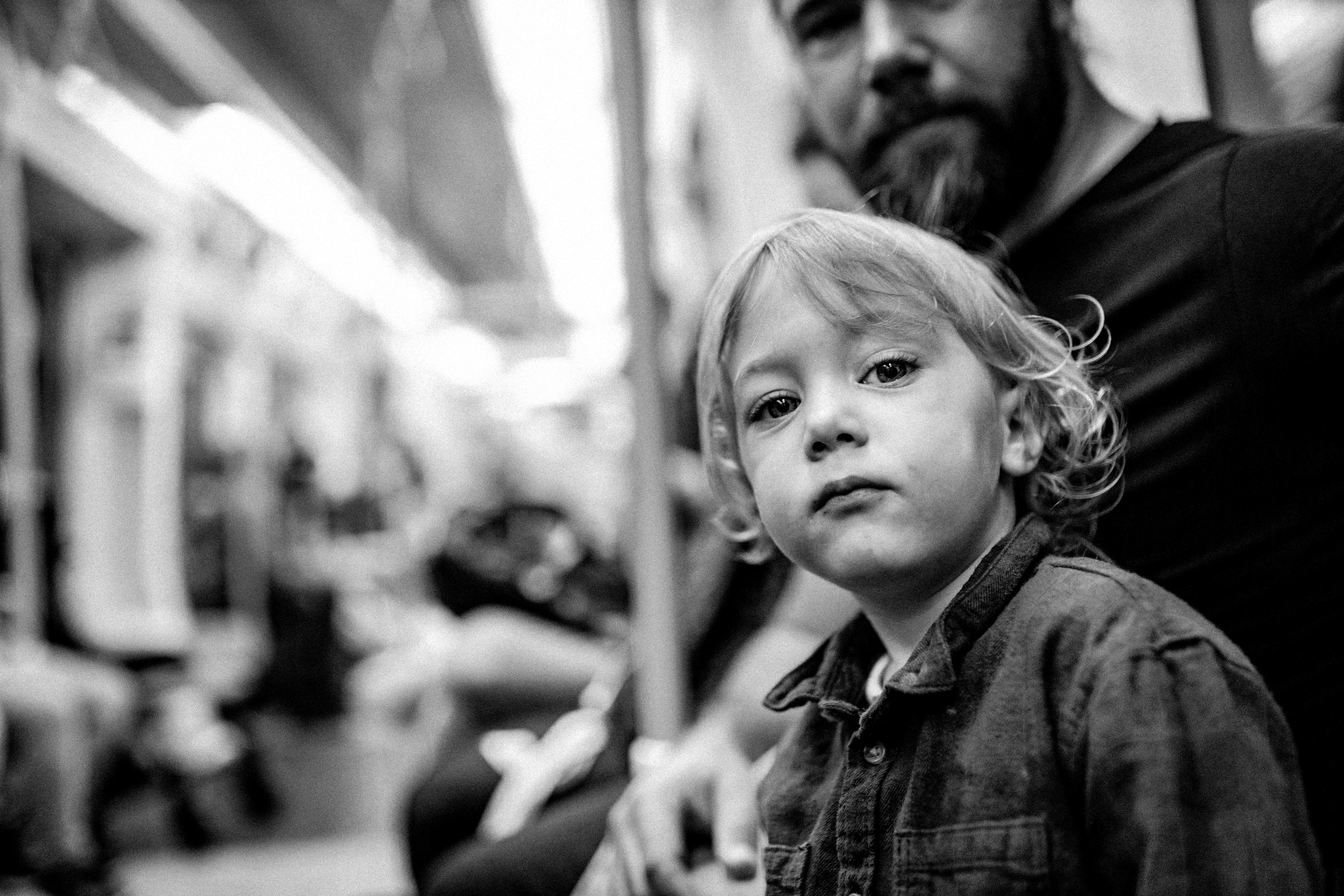 A young child looks toward the camera while riding public transport, an adult seated behind them.